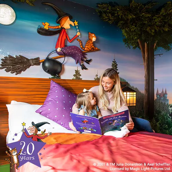 Child and woman sitting on bed reading Room on the Broom in a Room on the Broom Hotel room at the Chessington Resort Hotels celebrating 20 years of Room on the Broom